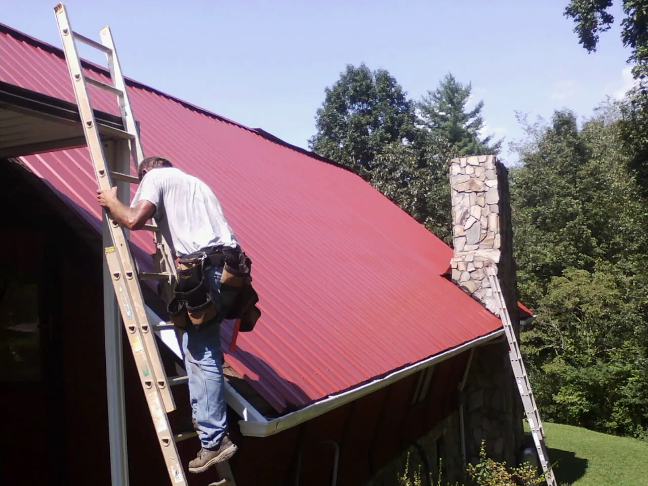 Roofer on a ladder beside a red metal roof and stone chimney — Soffit & Fascia Repair by Roofing Brothers in Bascom